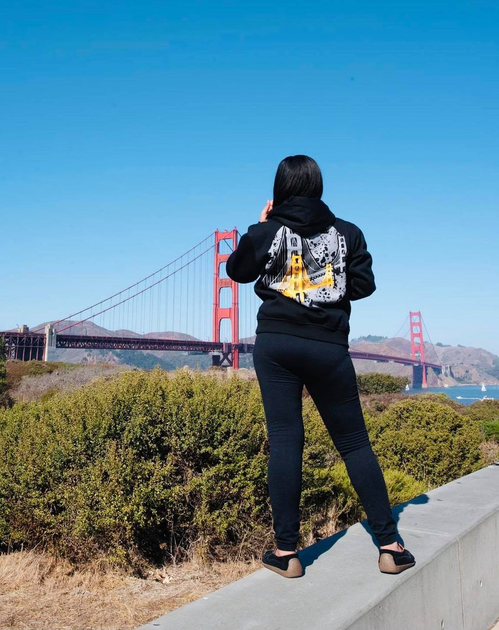 Hand printed Golden Gate Bridge Hoodie Black shown on  female model in front of the bridge.