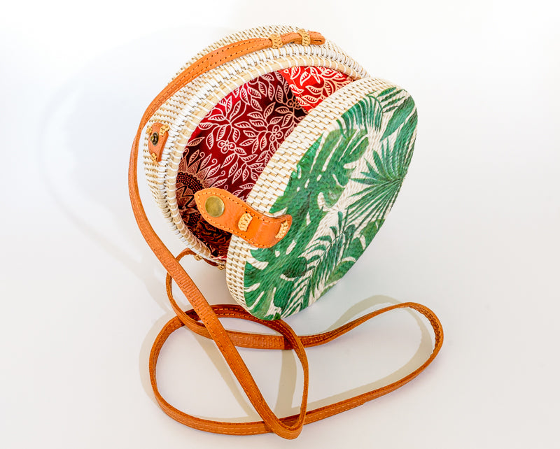 White rattan handbag hand woven in Bali Indonesia with tropical leaf decal, leather strap and brass clasp, showing colorful, patterned cotton lining.