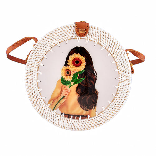 Round white rattan bag handwoven In Bali Indonesia, featuring painted wooden inlay of girl holding sunflowers, with leather strap & brass clasp.