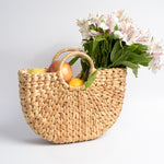 Roomy tote bag handmade in Bali from water hyacinth leaves, pictured to show sturdy overlapping weave with flat bottom and rounded sides. Sturdy round handles are woven directly into body.  Light tan in color.  Shown carrying fruit & flower bouquet.