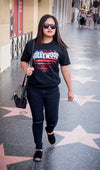 Black T-shirt front side showing the Hollywood Sign imposed over an American Flag motif, hand printed in red, white & blue, pictured on female model walking on Hollywood Blvd.
